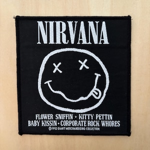 May include: A square black fabric patch featuring white embroidered text and graphics. The word NIRVANA appears at the top. Below is a circular smiley face with X eyes and a tongue sticking out. The bottom text reads FLOWER SNIFFIN KITTY PETTIN BABY KISSIN CORPORATE ROCK WHORES and 1992 GIANT MERCHANDISING COLLECTION.