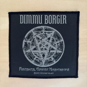 Può includere: Una toppa quadrata in tessuto nero con la scritta grigia DIMMU BORGIR in alto e PURITANICAL EUPHORIC MISANTHROPIA in basso. Al centro è presente un grafico circolare grigio di un pentagramma. Il testo ©2001 NUCLEAR BLAST è stampato sotto.