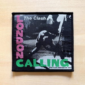 Puede incluir: Parche cuadrado negro con las palabras "LONDON CALLING" en rosa y verde, y "The Clash" arriba. La imagen muestra una silueta. El parche tiene un borde negro y está sobre una superficie de madera clara.