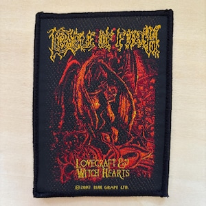 Puede incluir: Parche rectangular negro con el texto "Cradle of Filth" en amarillo, sobre una ilustración roja y naranja. El texto "Lovecraft & Witch Hearts" está debajo de la ilustración. La parte inferior del parche tiene el texto "©2005 BLUE GRAPE LTD."