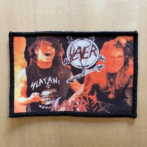 Puede incluir: Un parche rectangular de tela con un estampado gráfico del grupo Slayer. El diseño incluye a dos miembros de la banda, el logotipo de Slayer en un círculo blanco y la palabra Slayer en texto rojo en la parte inferior, con un borde negro cosido.