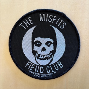 Puede incluir: Un parche bordado negro de forma circular con un gráfico blanco del logotipo de la calavera de Misfits en el centro. El texto THE MISFITS está arqueado arriba y FIEND CLUB abajo. Un pequeño texto en la parte inferior dice TM & © 2002 CYCLOPIAN MUSIC INC.