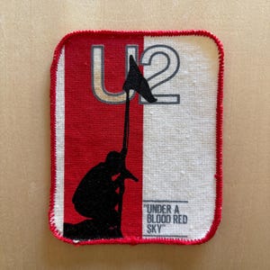 Könnte beinhalten: Ein rechteckiger Stoffaufnäher mit rotem und cremefarbenem Hintergrund. Er zeigt den Text U2 oben, eine schwarze Silhouette einer Person, die eine Flagge hält, und den Text UNDER A BLOOD RED SKY unten rechts, alles mit einem roten Rand.