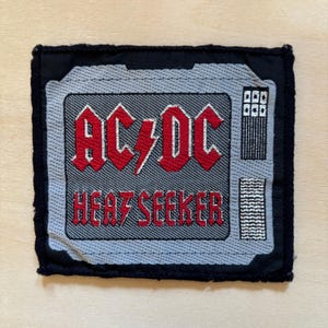 Op de afbeelding: Een vierkante geborduurde patch met een zwarte rand. Het midden toont een grijs televisieschermontwerp met de rode tekst AC/DC en HEAT SEEKER. Een rode bliksemschicht scheidt de letters AC en DC. Twee kleine luidsprekerroosters zijn zichtbaar.