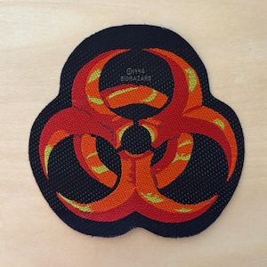 Puede incluir: Un parche bordado negro que presenta un símbolo de riesgo biológico rojo y naranja con detalles en amarillo. El texto "©1994 BIOHAZARD" está cosido en el centro del diseño. El parche tiene una forma redondeada de tres lóbulos con un borde de tela oscuro.
