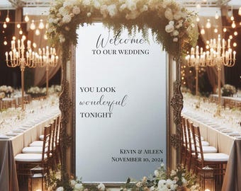 Op de afbeelding: Een grote, sierlijke spiegel met een gouden lijst die dient als welkomstbord voor een bruiloft. De tekst luidt: Welcome TO OUR WEDDING, YOU LOOK wonderful TONIGHT, KEVIN & AILEEN, NOVEMBER 10, 2024. De spiegel is versierd met witte bloemen.