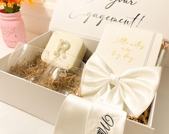 May include: A white gift box with the text Congratulations On Your Engagement containing two clear glasses, a square jewelry case with a pearl letter R, a notebook with the text Little notes for the big day, a white satin bow, and a white sash with Mrs. Bragga.