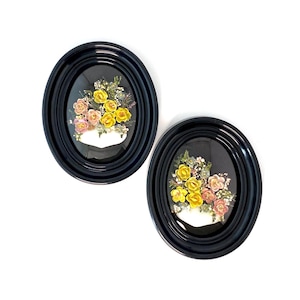 May include: Two oval framed pictures with black frames and glass fronts. Each frame contains a pressed flower arrangement with yellow, pink, and white flowers.