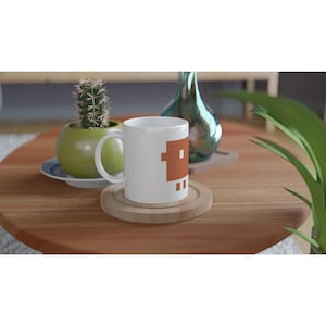 Könnte beinhalten: Weiße Keramiktasse mit braunem pixeligem Design, auf einem Holzuntersetzer. Ein kleiner Kaktus in einem grünen Topf und eine dekorative Glasvase befinden sich im Hintergrund. Die Tasse steht auf einem Holztisch.