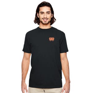 May include: Black t-shirt featuring a small, pixelated brown couch design on the upper left chest. The shirt is plain black and has short sleeves. The person wearing the shirt is smiling.