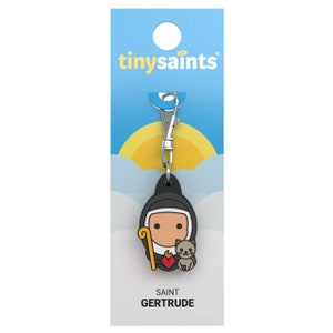 May include: A black and white cartoon keychain featuring Saint Gertrude, a nun with a cat and a heart. The keychain is attached to a silver clip and has the text "tiny saints" and "SAINT GERTRUDE" on the packaging.
