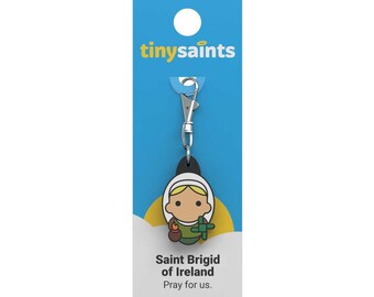 Puede incluir: Una tarjeta azul y amarilla con el texto "tinysaint" en ella. Un llavero plateado con una imagen de dibujos animados en negro y amarillo de Santa Brígida de Irlanda. El texto "Saint Brigid of Ireland" y "Pray for us." está en la tarjeta.