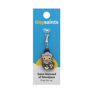 May include: A blue and yellow card with the text "tinysaint" on it. A silver keychain with a small, cartoon-style figure of Saint Bernard of Montjoux holding a dog. The text "Saint Bernard of Montjoux" and "Pray for us." is printed on the card.