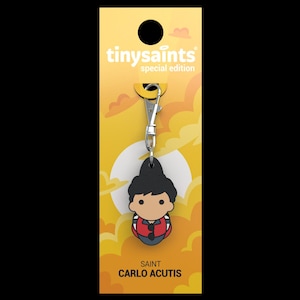 May include: A keychain featuring a cartoon illustration of Saint Carlo Acutis, a young man with black hair wearing a red shirt and a backpack. The keychain is part of the "tinysaint" special edition collection.