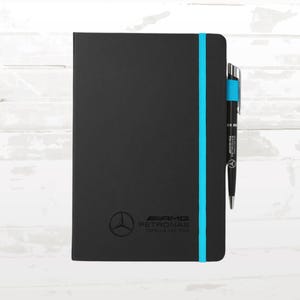 May include: Black notebook with a teal elastic band and a matching pen. The notebook features the AMG Petronas logo. The pen has teal accents and the Mercedes-Benz logo. The notebook and pen are on a white background.