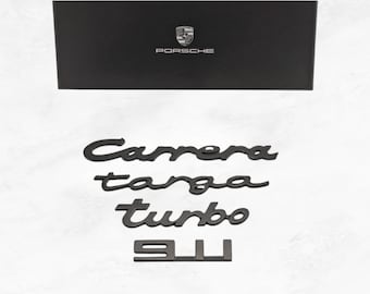 Puede incluir: Una caja rectangular negra con el logotipo y el texto de Porsche, acompañada de cuatro emblemas individuales de metal negro con las palabras Carrera, targa, turbo y el número 911 en una fuente cursiva y de bloque, dispuestos verticalmente.