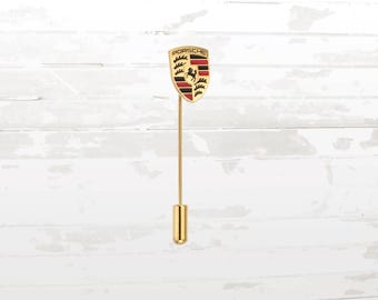 May include: A gold-colored lapel pin featuring the Porsche crest. The crest is a shield shape with black, red, and gold details, and the word "PORSCHE" is at the top. The pin has a long, slender stem and a rounded end.