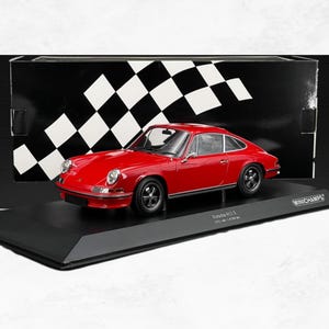 Peut inclure: Une voiture miniature Porsche 911 S rouge présentée sur un socle noir avec un fond à damier. Le modèle présente des roues noires, des vitres transparentes et des garnitures argentées. Le socle porte le texte Minichamps et Porsche 911 S 1973 red 1 of 504 pcs.
