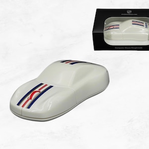 May include: A white computer mouse shaped like a sports car, featuring a red, white, and blue racing stripe down the centre. The mouse includes a red scroll wheel and is displayed alongside its black retail box with the Porsche logo and text.