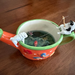 May include: A decorative watering can with a vibrant orange body, green spout and handle, and a white interior. The can is adorned with floral and butterfly designs. Miniature figurines of a white pug and a black and white cat fishing are perched on the can.