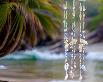May include: A seashell wind chime with blue, green, and white beads hanging from a wooden base. The wind chime is hanging in front of a blurred background of a beach and ocean.