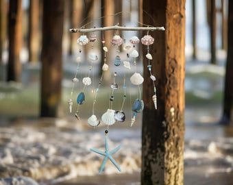 May include: A coastal-themed mobile featuring various seashells, beads, and a light blue starfish. The seashells are of different shapes and sizes, strung together with beads and suspended from a natural wooden branch. The mobile hangs in front of a blurred background of wooden pier posts and ocean waves.