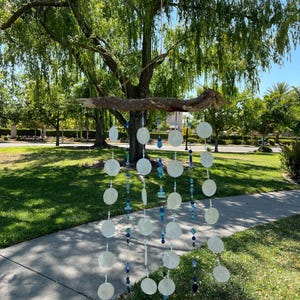 May include: A handcrafted wind chime made of natural wood and white, round shell discs. The chime features strands of blue and clear beads, creating a decorative and soothing outdoor accent. The wind chime hangs from a tree branch.