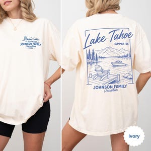 以下が含まれることがあります： 青いグラフィックデザインのアイボリー色のTシャツ。前面には、山のシーンの上に「JOHNSON FAMILY Vacation - 2026」というテキストがあります。背面には、湖のシーン、ボート、テキスト「JOHNSON FAMILY Vacation」とともに「Lake Tahoe SUMMER '26」が表示されています。