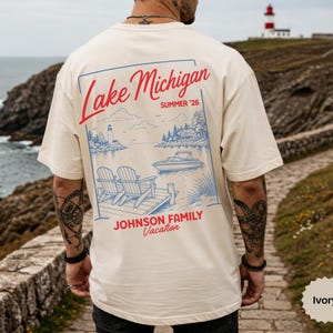 Puede incluir: Una camiseta de color marfil con un gr&aacute;fico azul y rojo en la espalda. El dise&ntilde;o incluye el texto Lake Michigan, Summer '26 y Johnson Family Vacation, con una ilustraci&oacute;n de dos sillas en un muelle, un barco y un faro.
