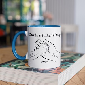 May include: White ceramic mug with a blue rim. The mug features a black and white illustration of two hands holding each other. The text "Our first Father's Day" is printed above the illustration. The text "Liam" and "Dad" are printed below the illustration. The year "2025" is printed below the text.