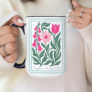 May include: White ceramic mug with a black handle and rim, featuring a floral design in pink and green. The design includes a tulip, daisy, and bell-shaped flowers within a green border. The text "Foxglove Daisy Tulip" is at the bottom.