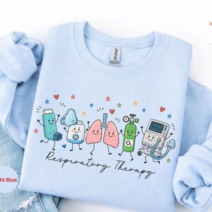 Puede incluir: Una sudadera de cuello redondo azul claro con un estampado gráfico de equipos médicos de dibujos animados que incluye un inhalador, nebulizador, pulmones, tanque de oxígeno y ventilador con el texto Respiratory Therapy en cursiva negra.