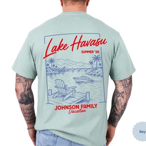 Puede incluir: Una camiseta verde claro con un gr&aacute;fico de l&iacute;neas azules en la espalda. El dise&ntilde;o muestra una escena de lago con una silla en un muelle, un barco y un puente. El texto rojo dice Lake Havasu, SUMMER '26 y JOHNSON FAMILY Vacation.