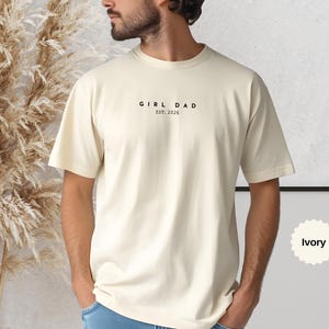 Pode incluir: Camiseta cor marfim com o texto preto "GIRL DAD EST. 2026". A camiseta, de gola redonda, &eacute; feita de um material macio. Sua cor neutra &eacute; adequada para todos.