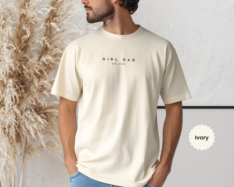 Pode incluir: Camiseta cor marfim com o texto preto "GIRL DAD EST. 2026". A camiseta, de gola redonda, &eacute; feita de um material macio. Sua cor neutra &eacute; adequada para todos.