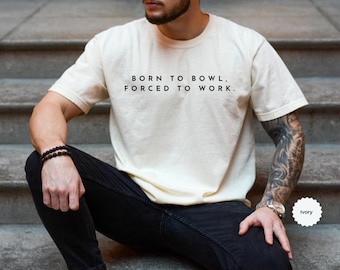May include: Ivory-colored t-shirt with the text "BORN TO BOWL, FORCED TO WORK." The shirt is a relaxed fit and is paired with black pants. A watch and bracelet are visible on the person's wrist.