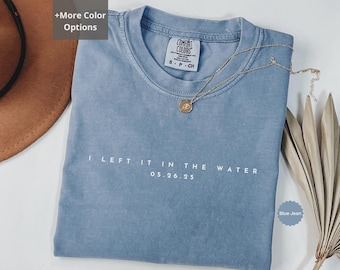 May include: A blue jean colored t-shirt with the text "I LEFT IT IN THE WATER 05.26.25" printed in white. A gold necklace with a pendant is draped across the shirt. The shirt is folded and placed on a surface with a brown hat and dried plant.