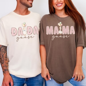 May include: A cream-colored t-shirt with the text DADA goose and a brown t-shirt with the text MAMA goose. Both shirts feature a graphic of a goose holding balloons, with the words DADA and MAMA in a pink plaid pattern and cursive text.