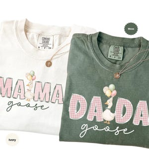 May include: Two short-sleeve t-shirts in ivory and moss green. The ivory shirt features the text MAMA goose with a goose wearing a party hat and balloons. The moss green shirt features the text DADA goose with a goose wearing a party hat and balloons. Both shirts have pink gingham lettering.