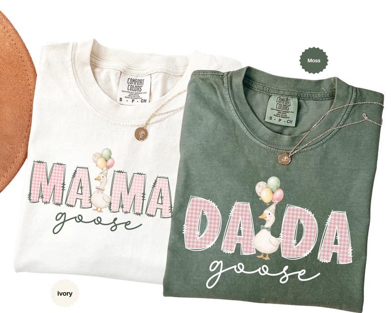 May include: Two short-sleeve t-shirts in ivory and moss green. The ivory shirt features the text MAMA goose with a goose wearing a party hat and balloons. The moss green shirt features the text DADA goose with a goose wearing a party hat and balloons. Both shirts have pink gingham lettering.