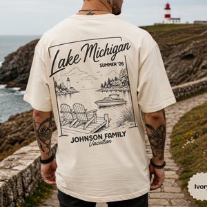 以下が含まれることがあります： アイボリー色のTシャツ。灯台、ボート、椅子のある桟橋など、湖のシーンの白黒グラフィックが描かれています。「Lake Michigan Summer '26」と「Johnson Family Vacation」の文字もプリントされています。