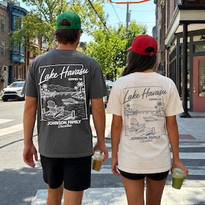 以下が含まれることがあります： 湖の風景が描かれたTシャツを着た2人が、通りを歩いています。Tシャツはグレーとクリーム色で、「Lake Havasu Summer '26」の文字とヤシの木のグラフィックがプリントされています。Tシャツには「Johnson Family Vacation」とも書かれています。