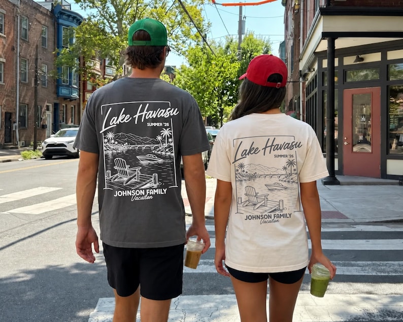 以下が含まれることがあります： 湖の風景が描かれたTシャツを着た2人が、通りを歩いています。Tシャツはグレーとクリーム色で、「Lake Havasu Summer '26」の文字とヤシの木のグラフィックがプリントされています。Tシャツには「Johnson Family Vacation」とも書かれています。
