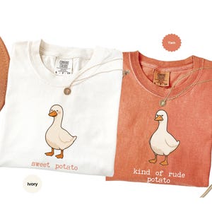 May include: Two Comfort Colors t-shirts, one ivory and one yam-colored, each featuring a cartoon duck illustration. The ivory shirt reads "sweet potato," while the yam shirt says "kind of rude potato." Both shirts have a gold necklace.