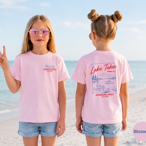 Puede incluir: Una camiseta de manga corta de color rosa claro con un peque&ntilde;o gr&aacute;fico en el pecho y un gr&aacute;fico grande en la espalda. El dise&ntilde;o posterior muestra una escena de lago con monta&ntilde;as, un barco y una silla, con el texto Lake Tahoe Summer 26.