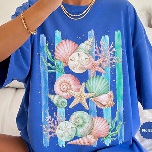 May include: A blue t-shirt featuring a watercolor design of seashells, starfish, and coral. The design includes pink, green, and white shells, with blue vertical stripes. The shirt has short sleeves and a relaxed fit.
