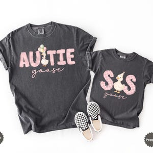May include: Two charcoal gray t-shirts featuring pink gingham lettering. The larger shirt reads "AUNTIE goose" with a goose holding balloons. The smaller shirt reads "SIS goose" with a goose wearing a party hat. A pair of black and white checkered slip-on shoes.
