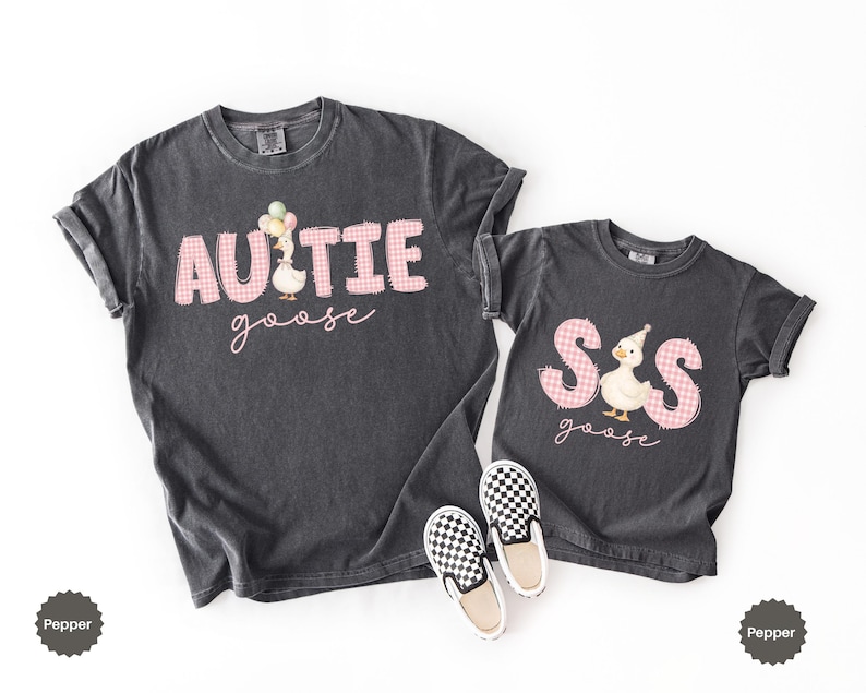 May include: Two charcoal gray t-shirts featuring pink gingham lettering. The larger shirt reads "AUNTIE goose" with a goose holding balloons. The smaller shirt reads "SIS goose" with a goose wearing a party hat. A pair of black and white checkered slip-on shoes.