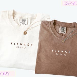 Puede incluir: Dos camisetas, una marfil y otra espresso, con la palabra "FIANC&Eacute;E" y la fecha "04.04.26" impresas en la parte delantera. Cada camiseta tiene cuello redondo y un peque&ntilde;o collar con colgante. Las camisetas est&aacute;n etiquetadas como "Comfort Colors".