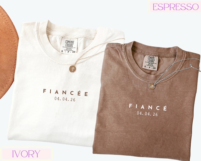 Puede incluir: Dos camisetas, una marfil y otra espresso, con la palabra "FIANC&Eacute;E" y la fecha "04.04.26" impresas en la parte delantera. Cada camiseta tiene cuello redondo y un peque&ntilde;o collar con colgante. Las camisetas est&aacute;n etiquetadas como "Comfort Colors".